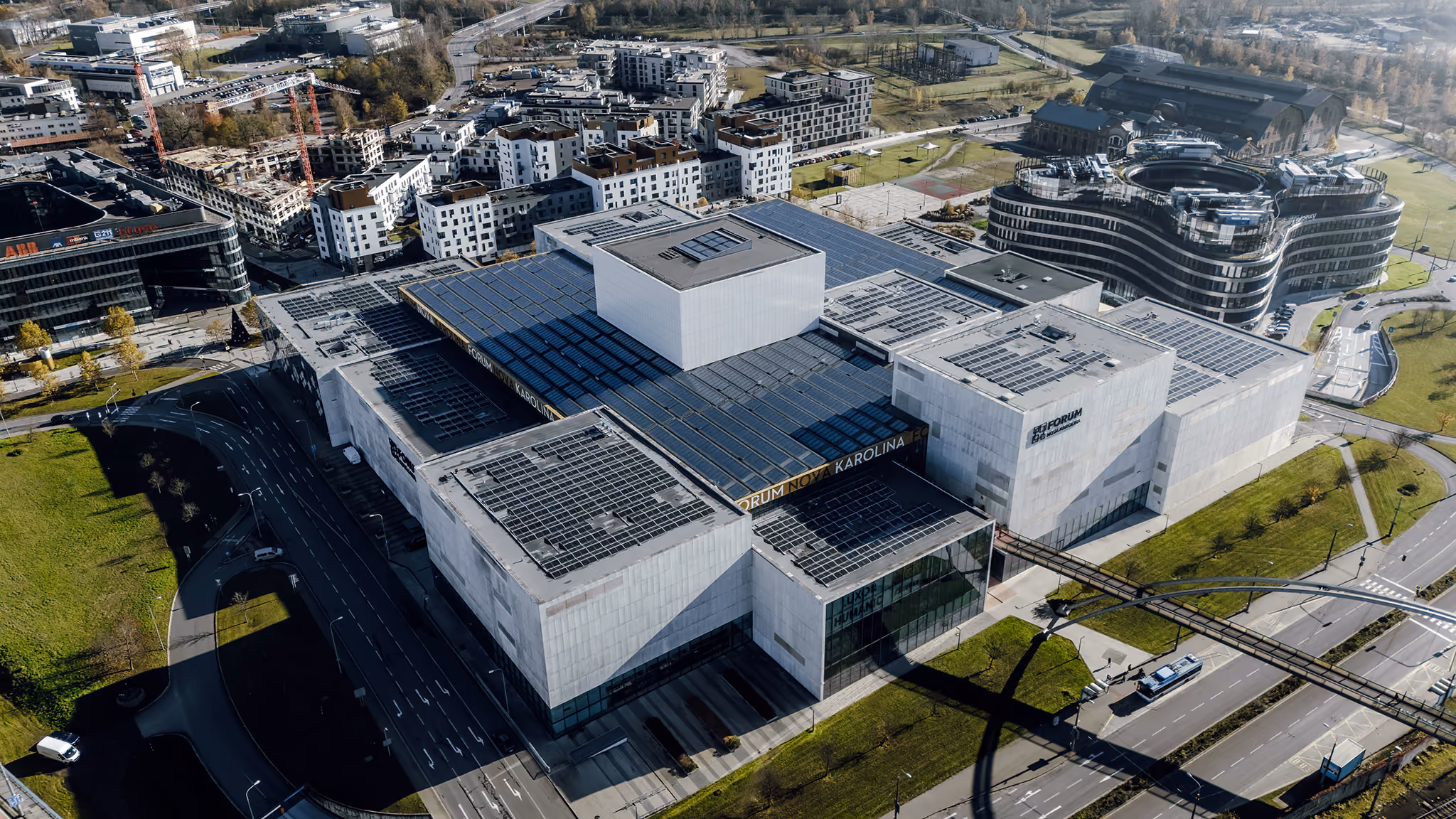 Aerial view of a large modern building with solar panels on its flat roof, surrounded by roads, green lawns, and residential and office buildings in an urban area.