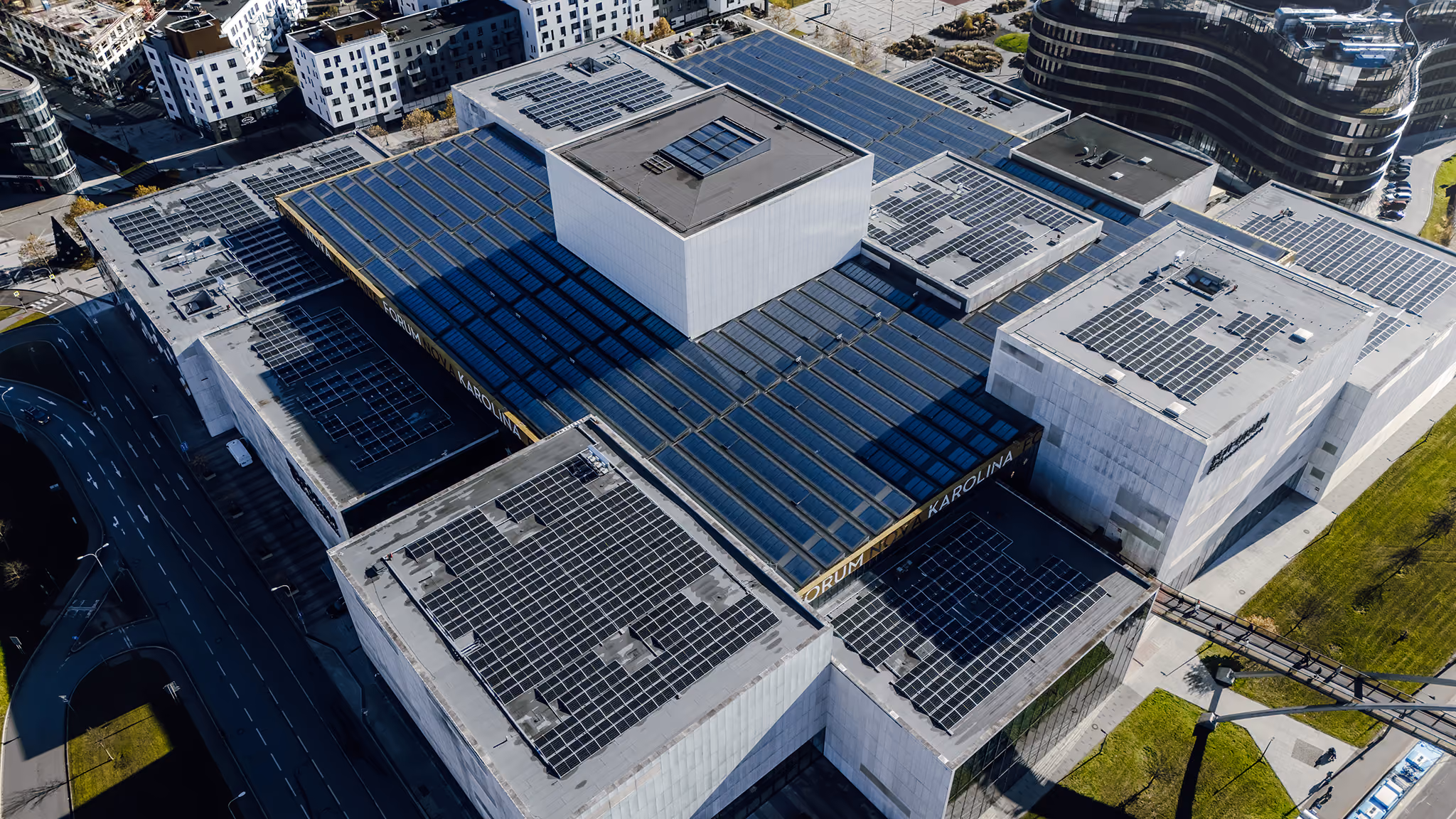 Aerial view of a modern urban complex with multiple large white buildings featuring flat roofs equipped with solar panels, connected by a skybridge, surrounded by roads and green areas.