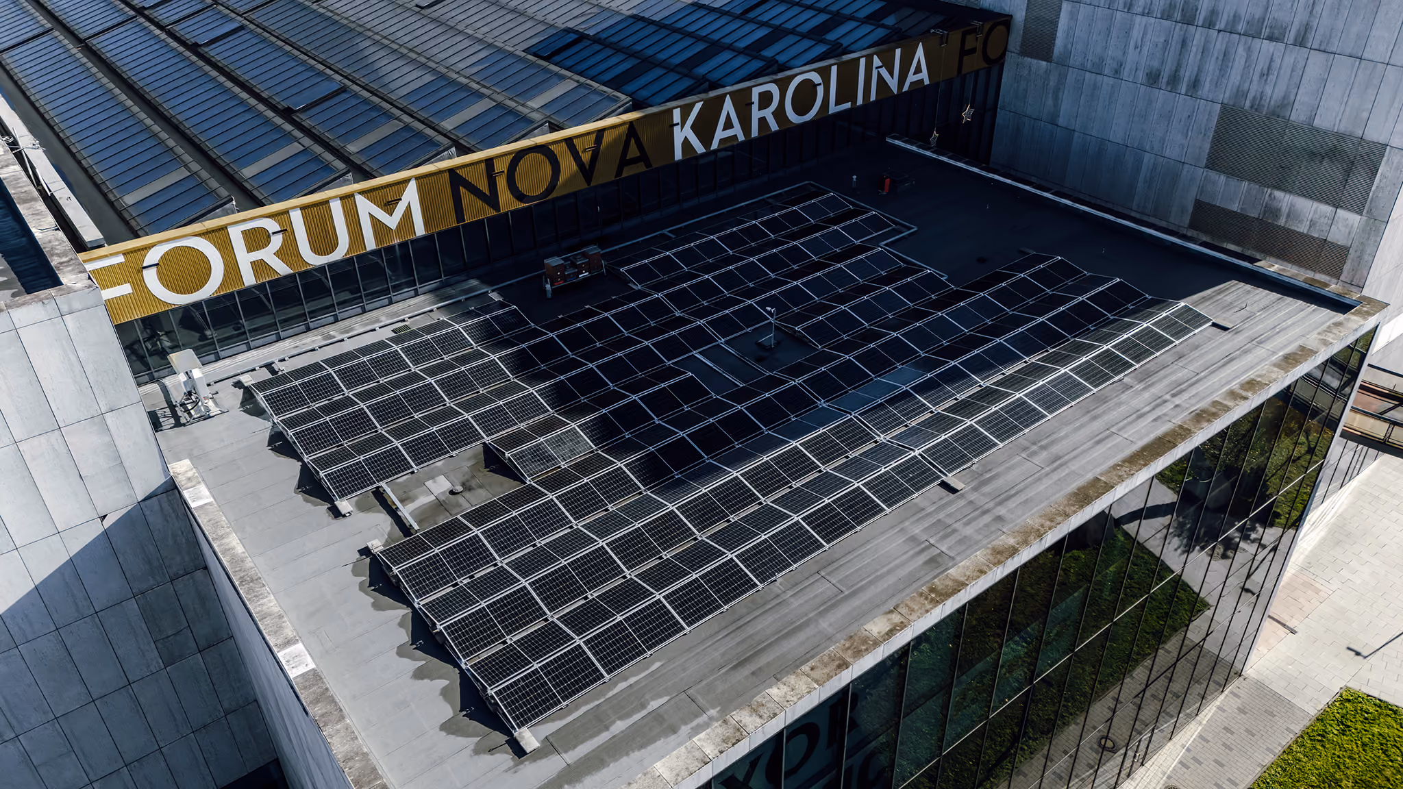 Aerial view of a building rooftop covered with multiple solar panels, with a sign reading 'FORUM NOVA KAROLINA' visible on the upper facade.