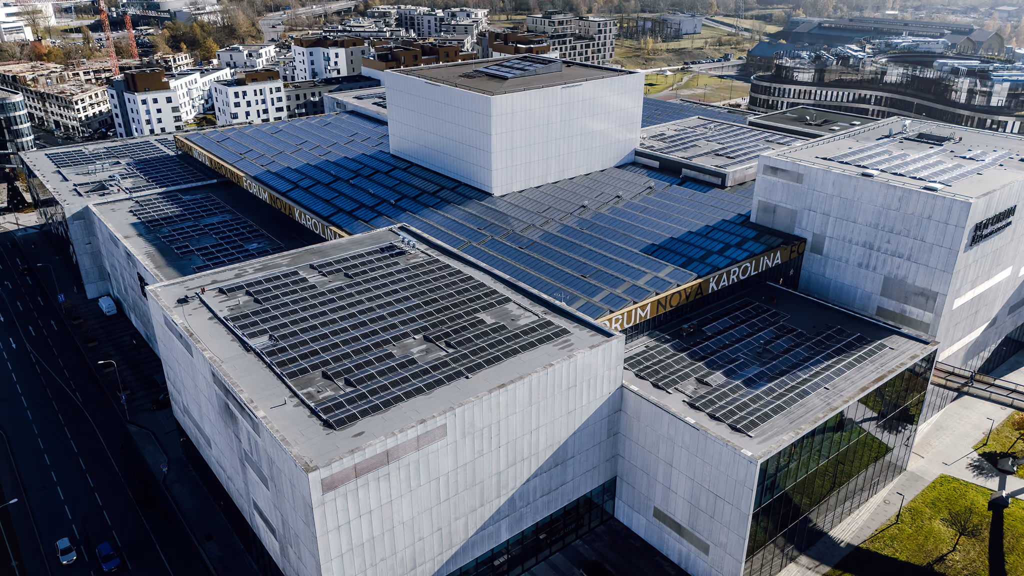 Aerial view of a modern, large white building complex with flat roofs covered in solar panels, surrounded by roads and other buildings.