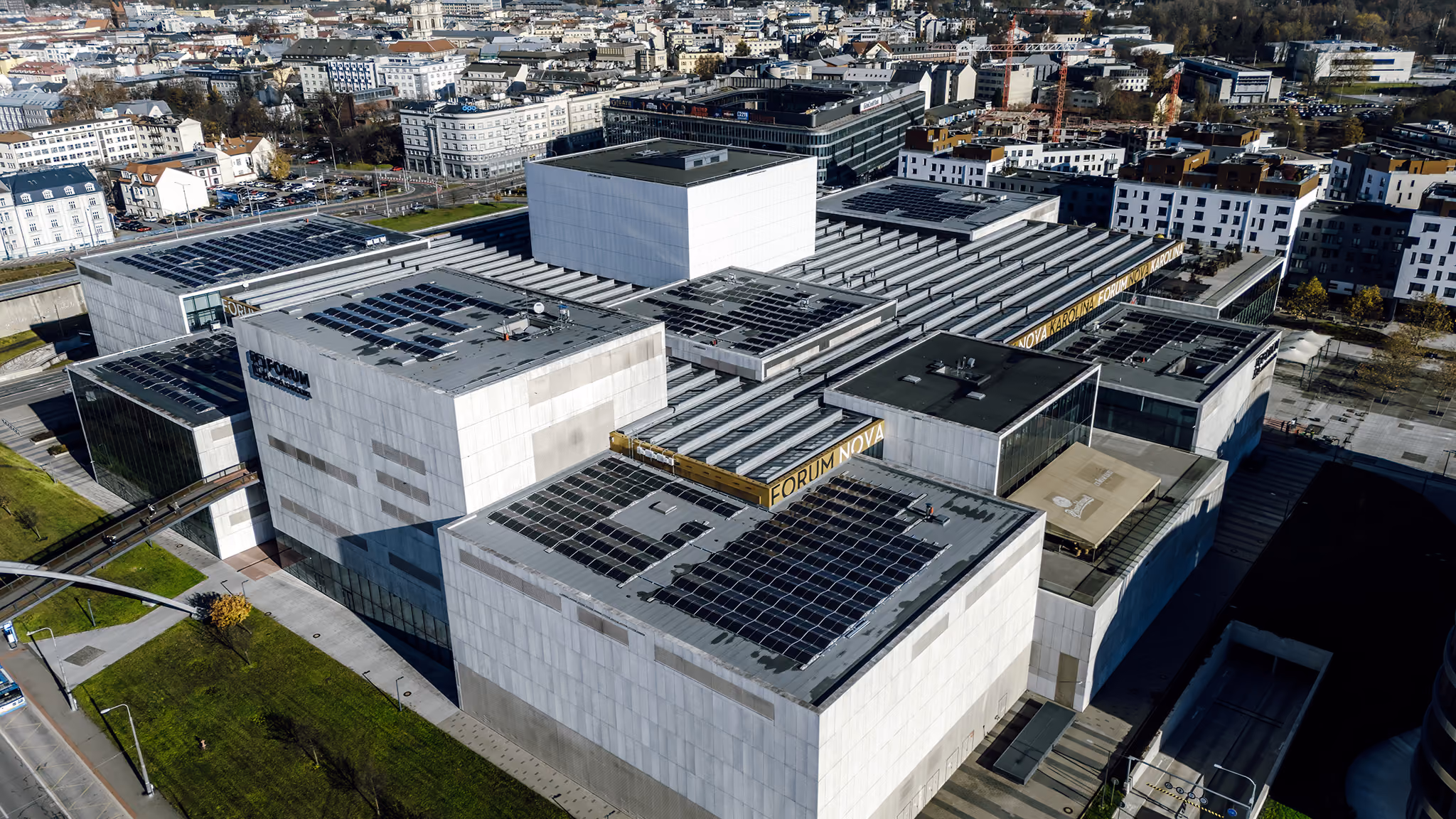 Aerial view of a modern, large building complex with flat roofs covered in solar panels, surrounded by urban cityscape and greenery.
