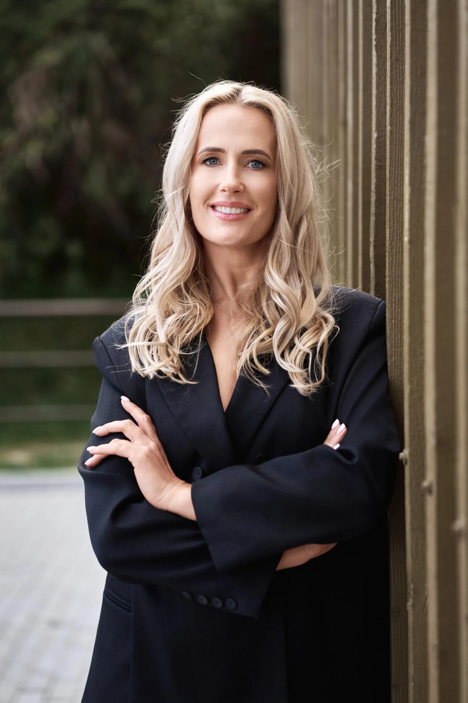 Moteris su ilgais blondinų plaukais, dėvinti juodą švarką, pasidėjusi rankas sukryžiuotas ir besišypsanti, remiasi į sieną.