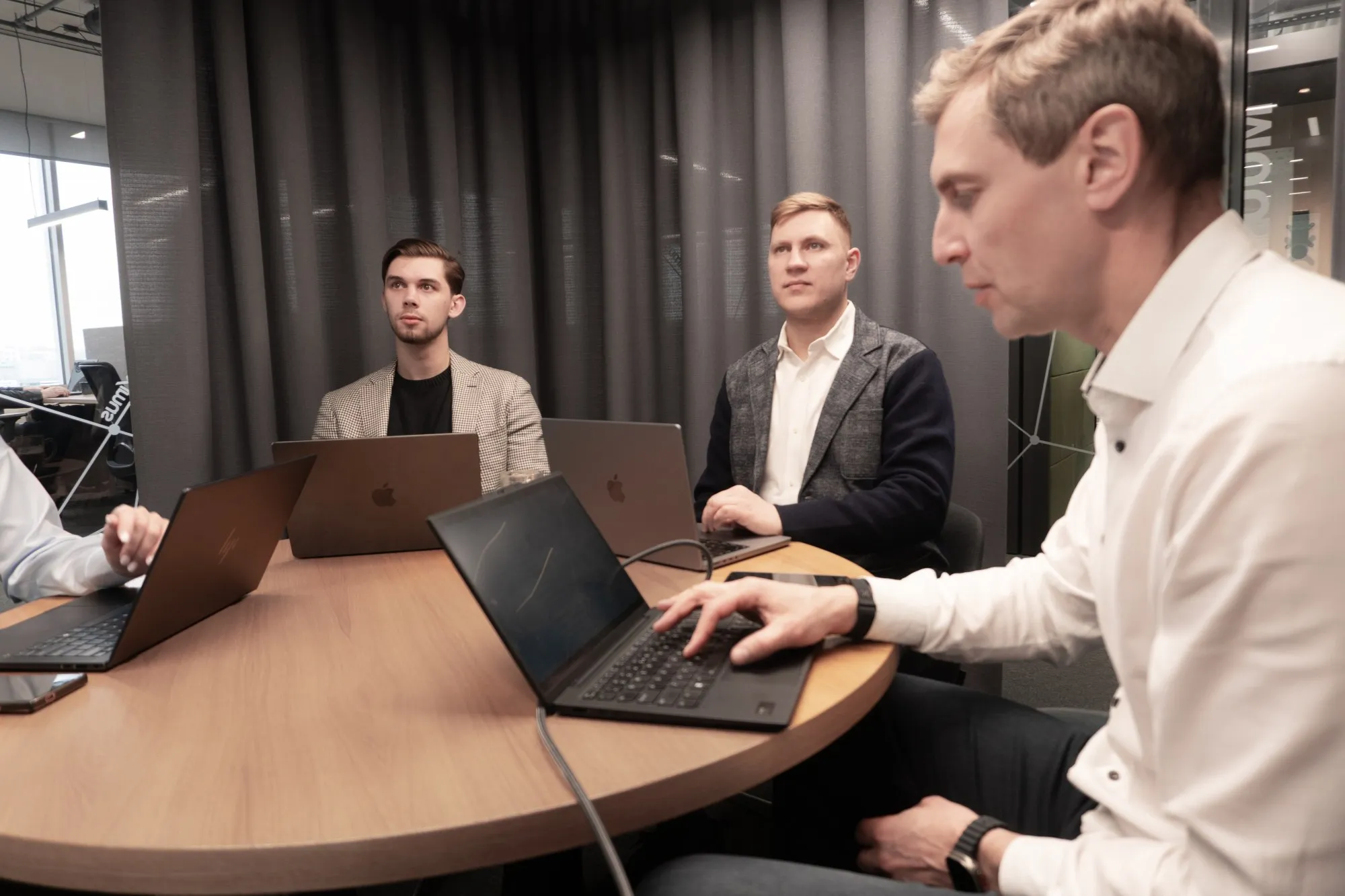 Keturi vyrai susirinkę prie apvalaus medinio stalo dirba su nešiojamaisiais kompiuteriais moderniame biure.