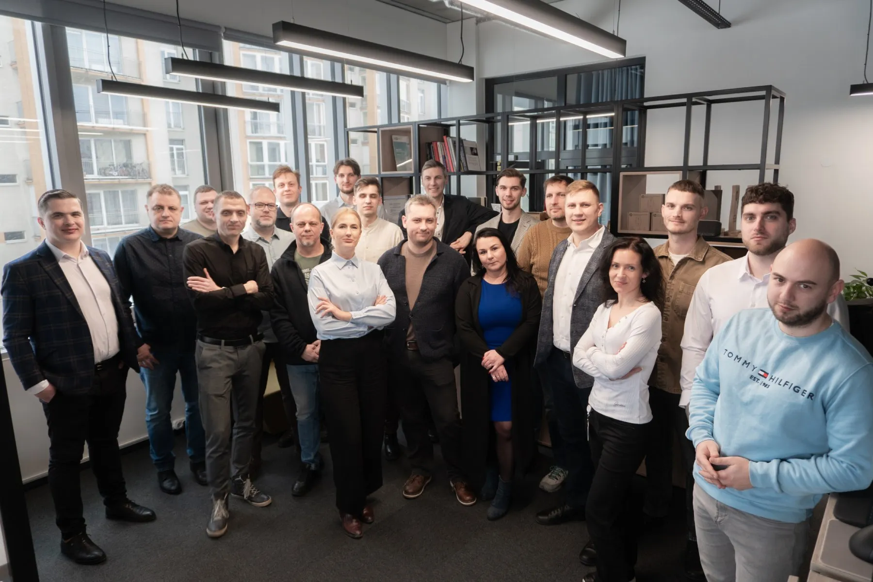 Dvidešimt žmonių grupė biuro patalpoje stovi ir žiūri į kamerą.