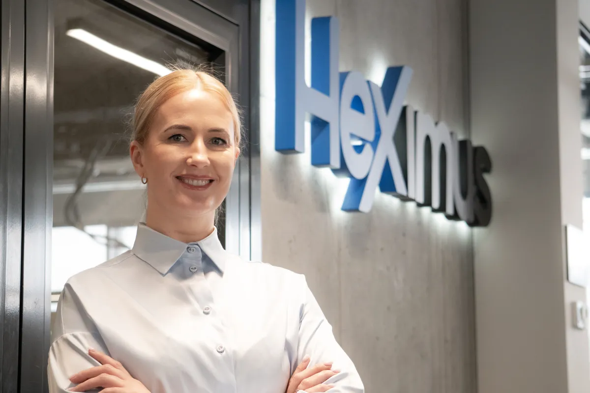 Šypsosi moteris baltu marškiniu, stovinti prie sienos su užrašu „Heximus“ biuro fone.