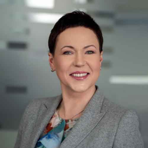 Portretas besišypsančios trumpaplaukės moters su pilka švarku ir spalvota palaidine neutralioje aplinkoje.