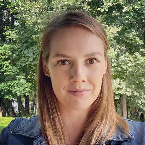 Moteris su šviesiais plaukais ir džinsine striuke, stovinti lauke prie žaliuojančių medžių.