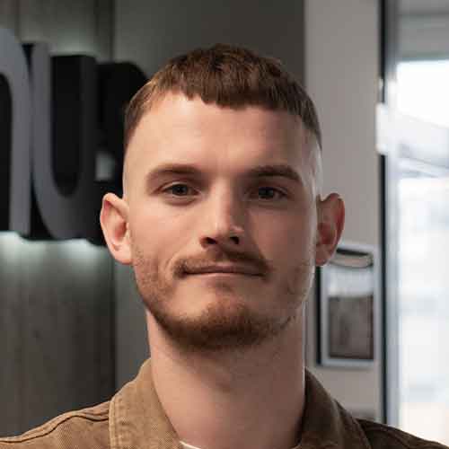 Jaunas vyras trumpais šviesiais plaukais ir barzda žvelgia į priekį viduje.