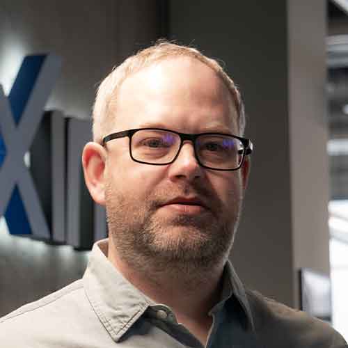 Vyras su akiniais stovi viduje, fone matoma pilka siena su iš dalies matomu mėlynu ir juodu užrašu.