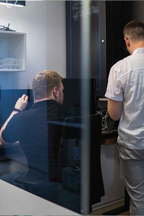 Du vyrai dirba biuro aplinkoje, vienas sėdi prie stalo, kitas stovi šalia.