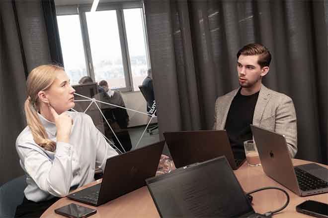 Du žmonės sėdi prie apvalaus stalo su nešiojamais kompiuteriais ir diskutuoja biure.