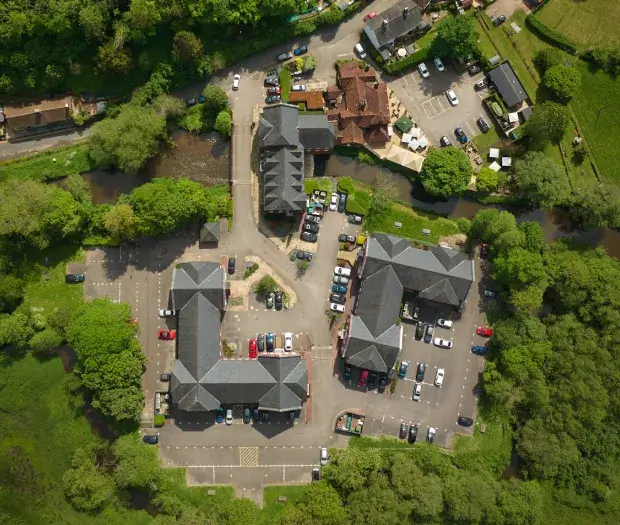 Aerial view of two large buildings with gray roofs surrounded by a parking lot filled with cars and lush green trees, next to a river.