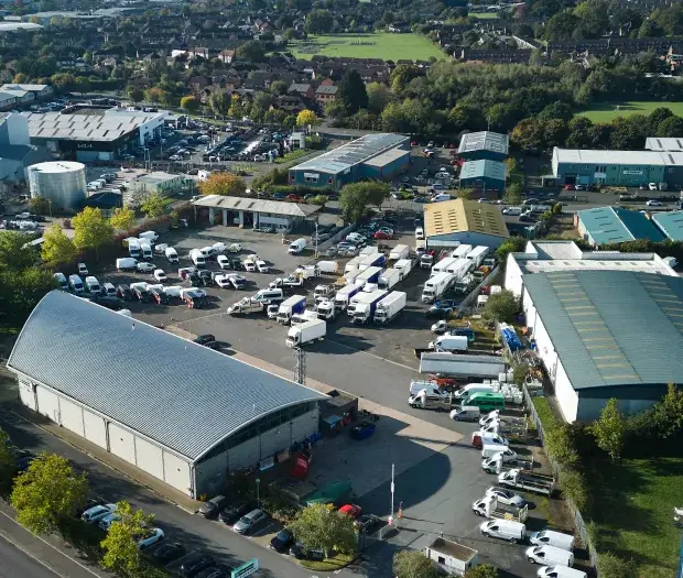 Aerial view of an industrial property with multiple warehouses, parked trucks, vans, and surrounding greenery.