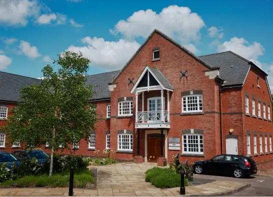 Red brick office building with white-trimmed windows, a central balcony, and cars parked nearby under a partly cloudy sky.