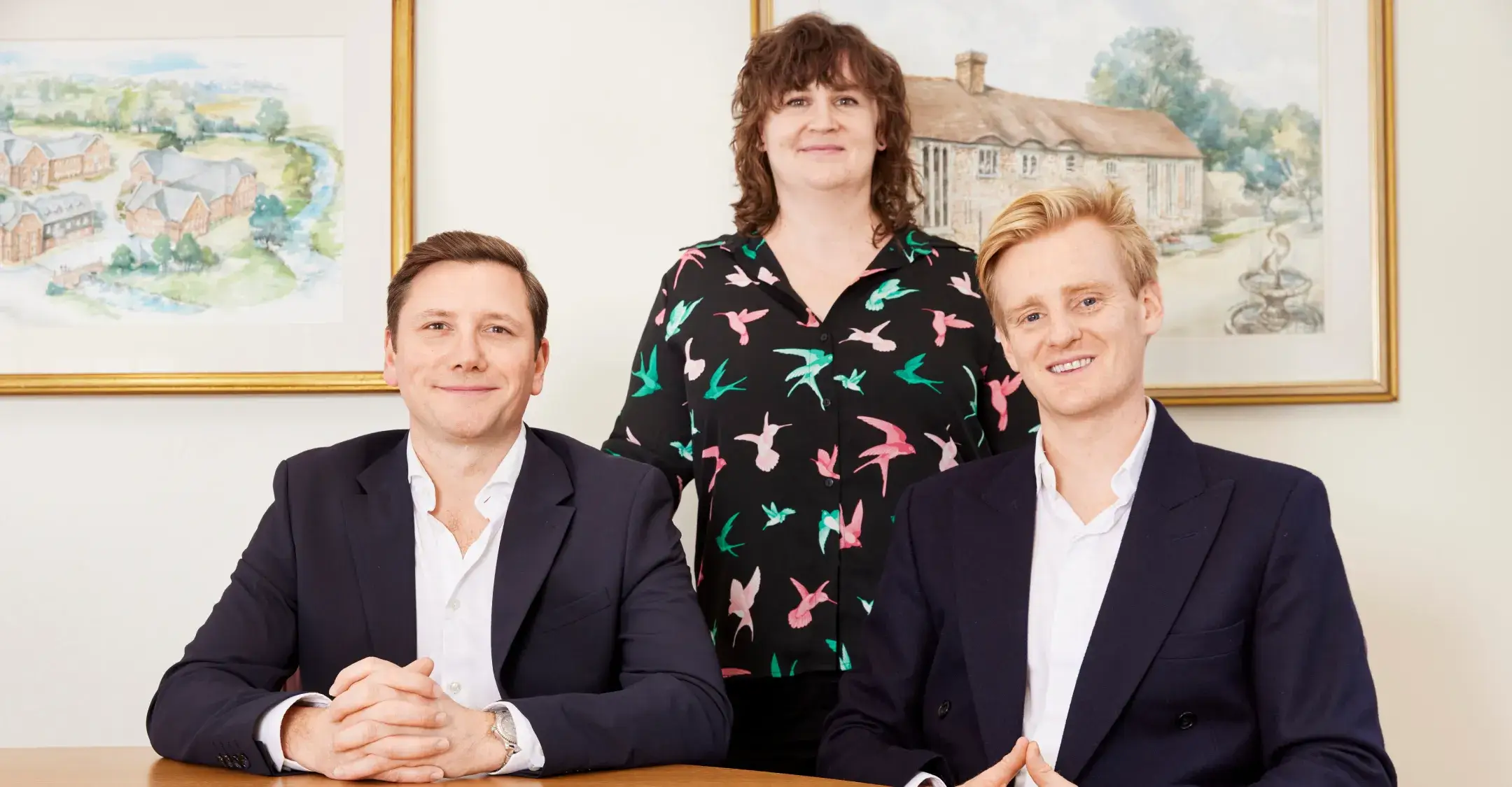 Two men in suits seated at a table with a woman in a bird-patterned shirt standing behind them, all smiling.