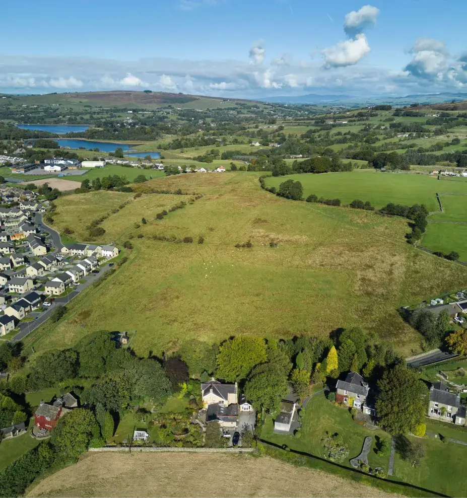 Aerial view of a rural landscape with scattered houses, green fields, and rolling hills under a partly cloudy blue sky.