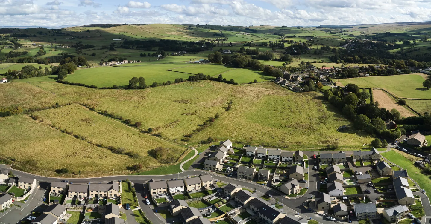 Aerial view of a residential neighborhood with houses and green fields extending into rolling hills under a partly cloudy sky.