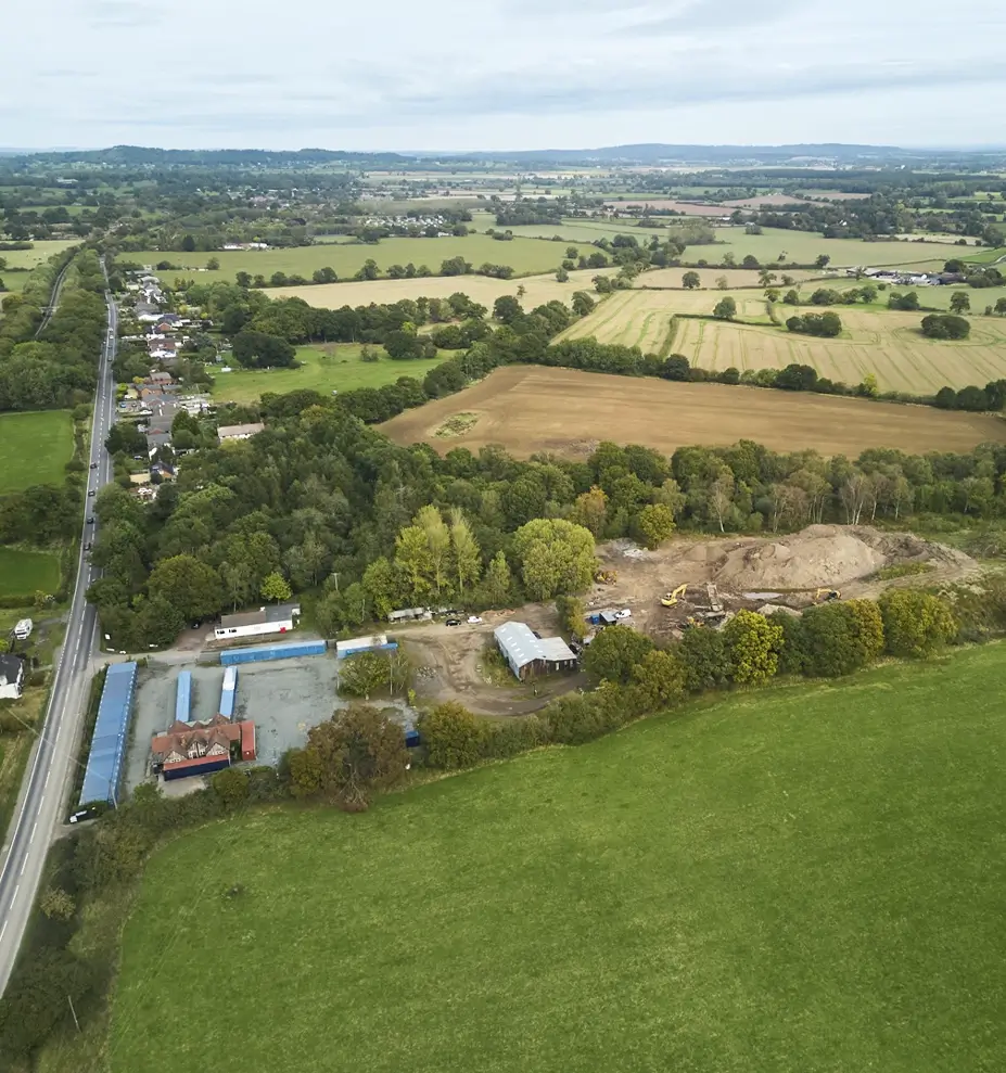 Aerial view of a rural landscape with green fields, farmland, patches of trees, a road, and a construction site with machinery.
