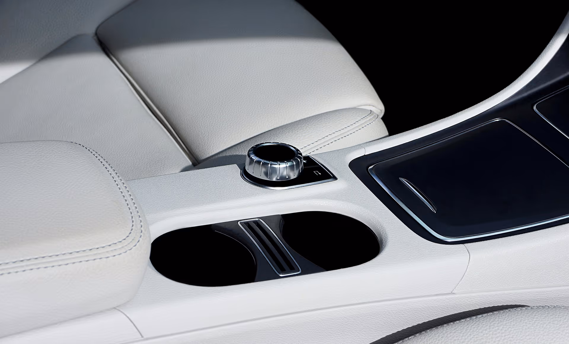 Close-up of a car interior featuring light gray leather seats, a rotary control knob, and dual cup holders with a black panel nearby.