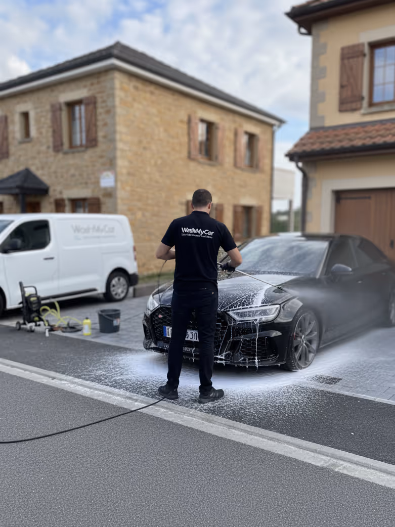 Un homme lave une voiture noire dans une allée devant une maison en pierre avec un van blanc garé à côté.