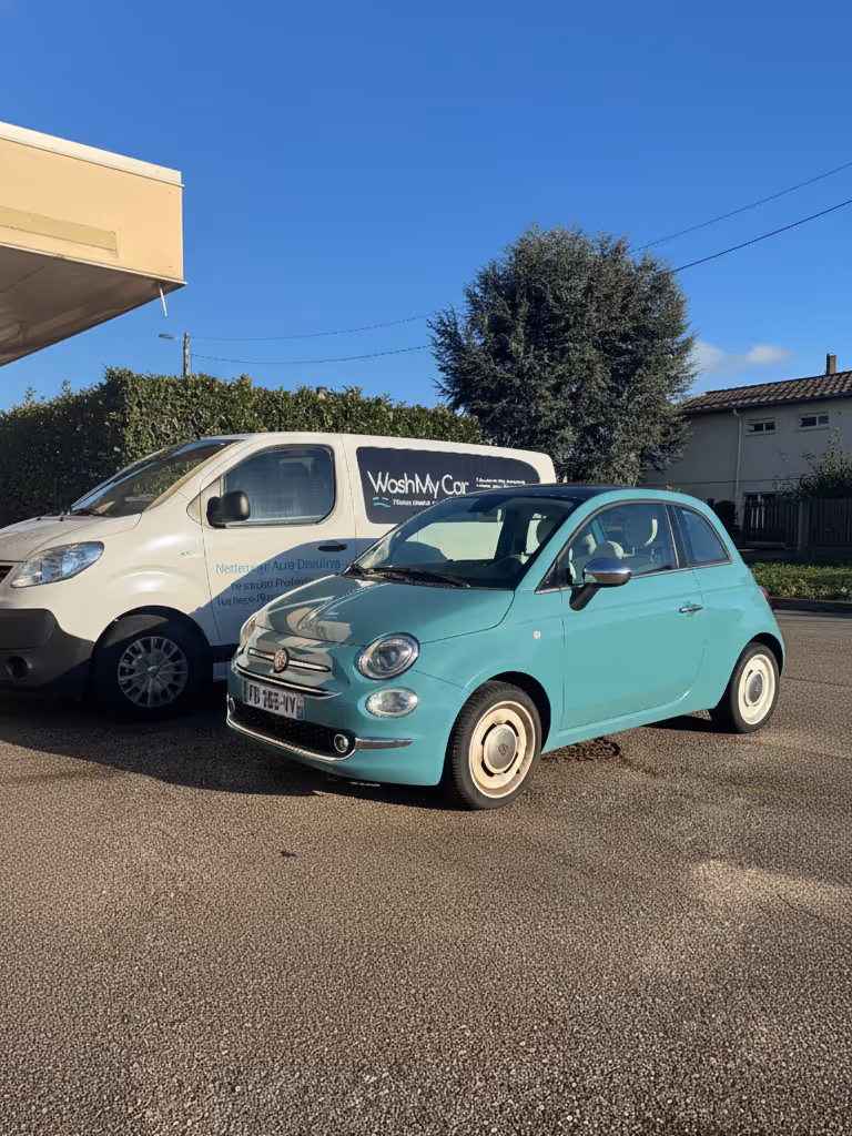 Petite voiture turquoise stationnée devant un utilitaire blanc avec le logo WashMyCar sous un ciel bleu clair.