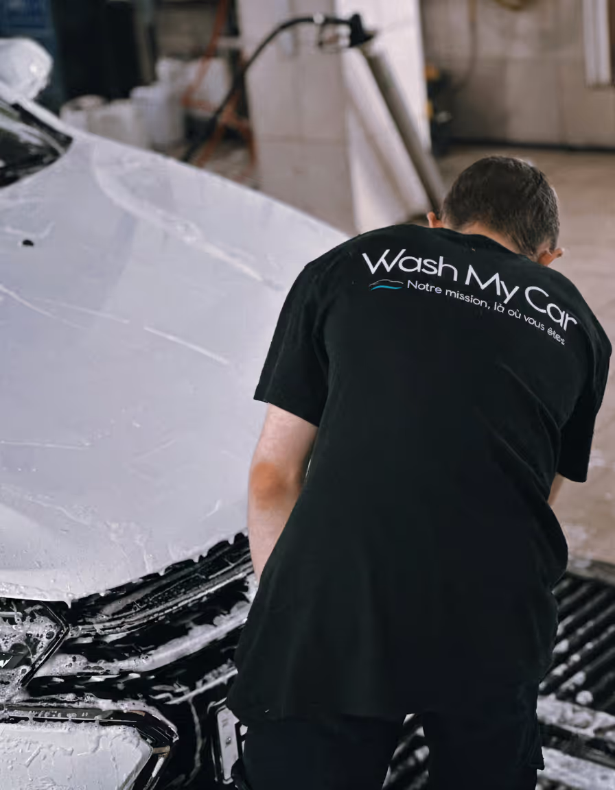 Homme de dos portant un t-shirt noir Wash My Car avec la phrase Notre mission, là où vous êtes, en train de laver une voiture blanche couverte de mousse.