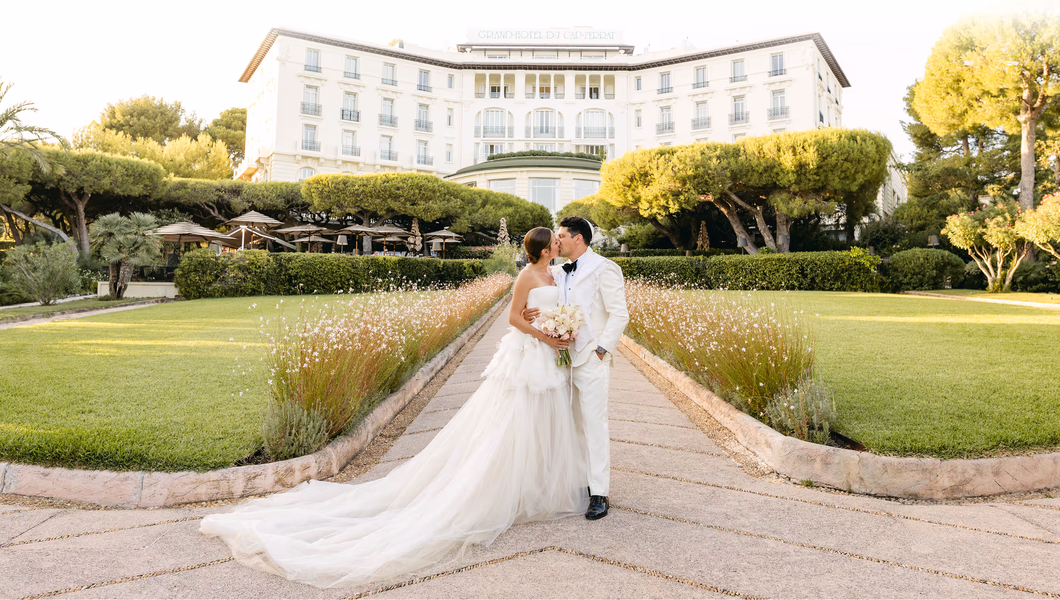 Bride and groom in white attire kissing on a paved garden path with a grand hotel and manicured greenery in the background.