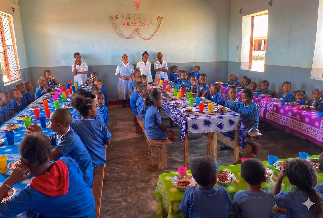 Une salle de réfectoire lumineuse à Madagascar où de nombreux enfants en uniforme bleu sont assis à de grandes tables colorées pour le repas.