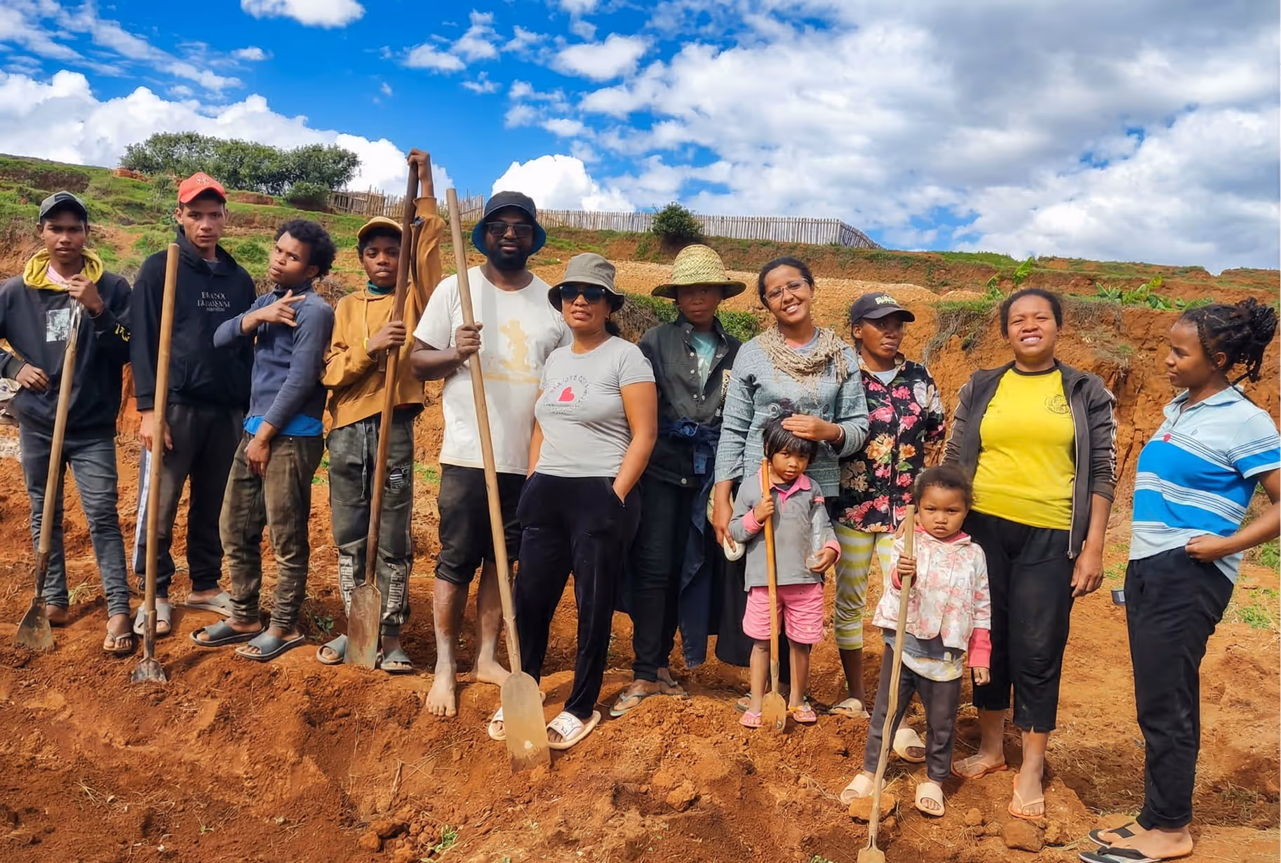 Une équipe multiculturelle et locale de travailleurs et d'habitants, composée de quinze personnes, pose fièrement sur un terrain agricole aménagé à Madagascar. Ils sont debout sur un sol rouge-brun, tenant des outils agricoles comme des pelles, des râteaux et des brouettes. 