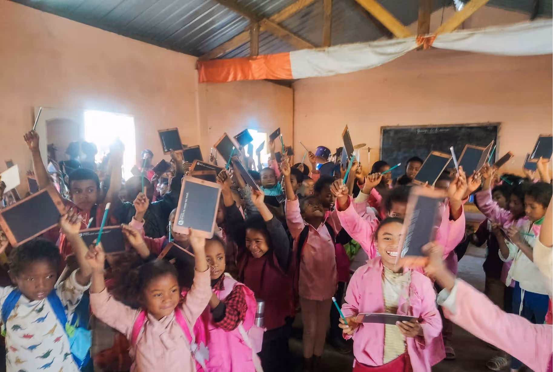 Un groupe joyeux d'écoliers et de professeurs malgaches dans une salle de classe. De nombreux enfants, vêtus d'uniformes, lèvent les bras avec enthousiasme, tenant de nouvelles ardoises d'écolier et des crayons. Au centre, une enseignante au grand sourire distribue une ardoise. La photo montre le matériel scolaire distribué à Madagascar.