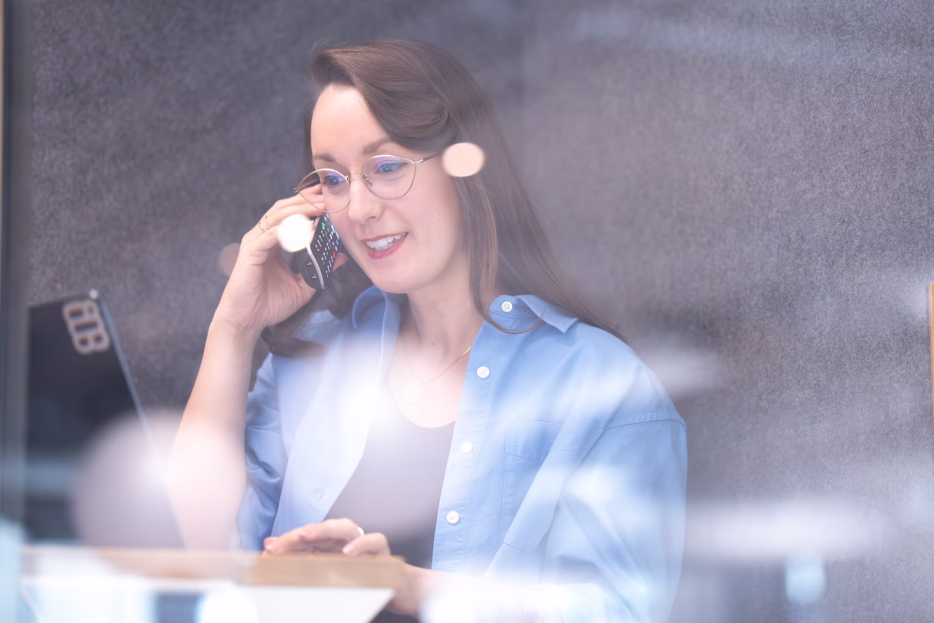 Frau mit Brille spricht lächelnd am Telefon in einem Büro mit Laptop vor sich.