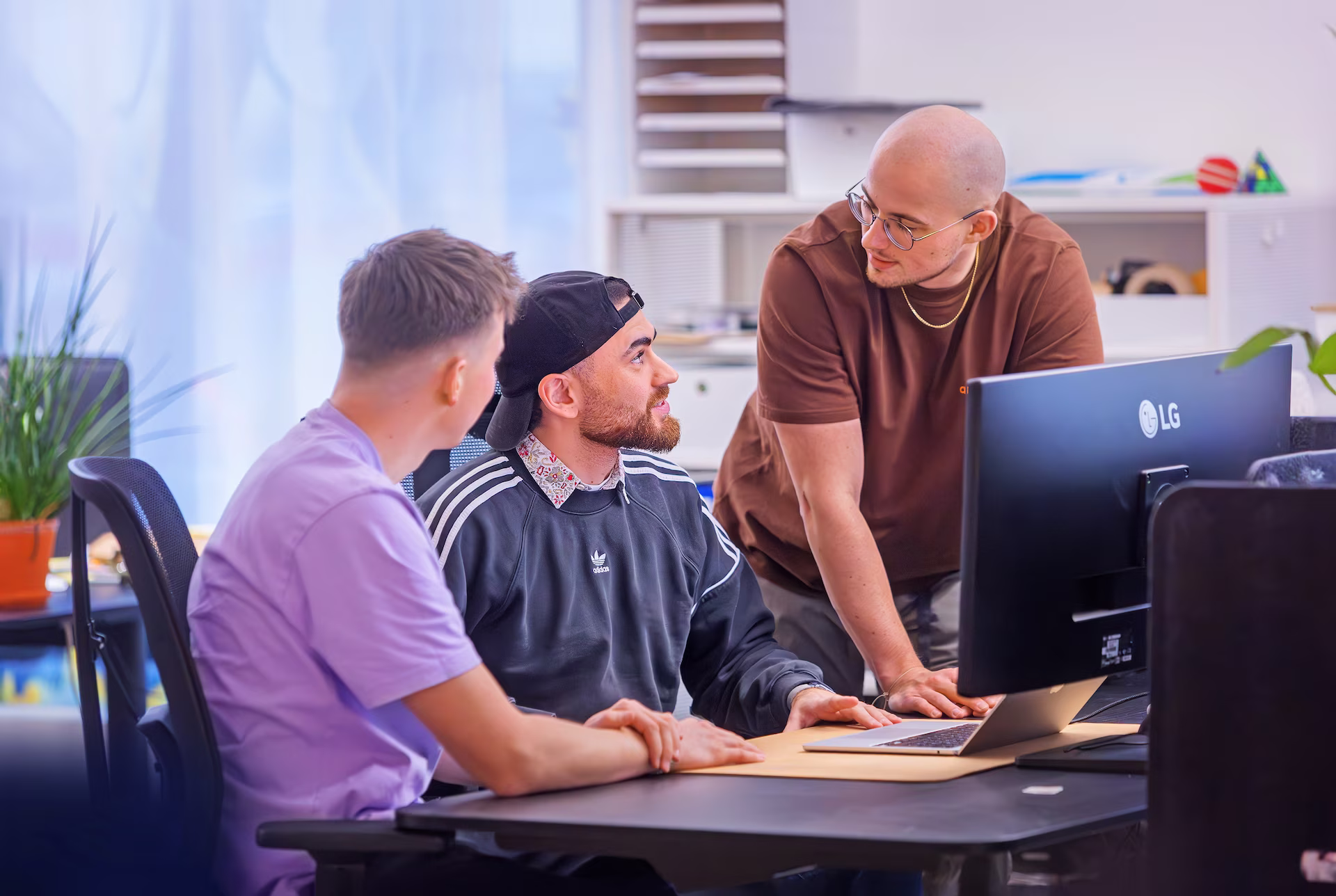 Drei Männer sitzen und stehen zusammen vor einem Computerbildschirm in einem modernen Büro und diskutieren.