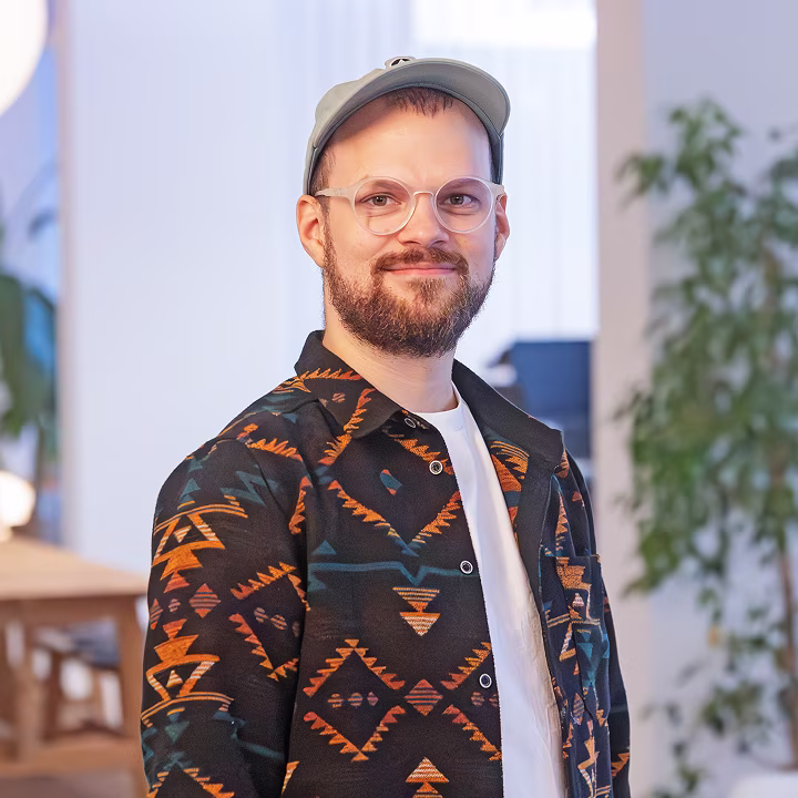 Porträt eines lächelnden Mannes mit Brille, Bart und Cap, der eine gemusterte Jacke trägt, vor einem Innenhintergrund mit Pflanzen.