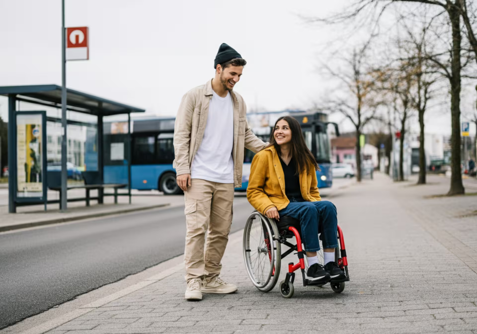 Mann steht lächelnd neben einer Frau im Rollstuhl an einer Bushaltestelle in der Stadt.