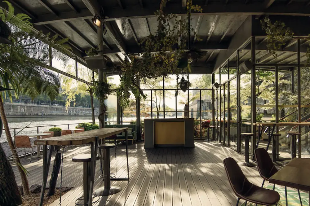 Modern outdoor café with wooden tables, assorted chairs, hanging plants, and river view through large glass windows.