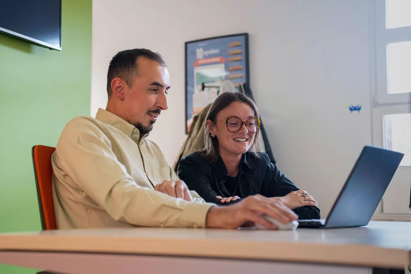 Due persone sedute a un tavolo sorridono mentre guardano lo schermo di un laptop in un ufficio.