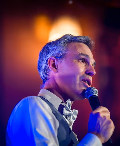 Man with gray hair wearing a bow tie and vest speaking into a microphone with warm blurred background lighting.