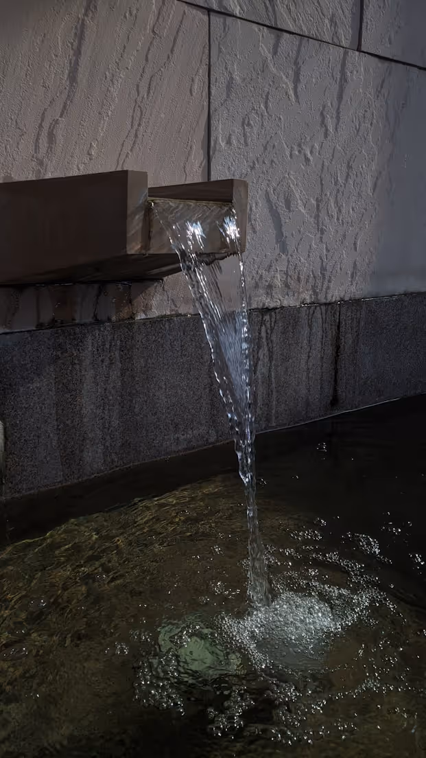 Water flowing from a rectangular metal spout into a shallow pool against a textured stone wall.