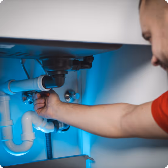 Person in a red shirt working on plumbing pipes under a sink.