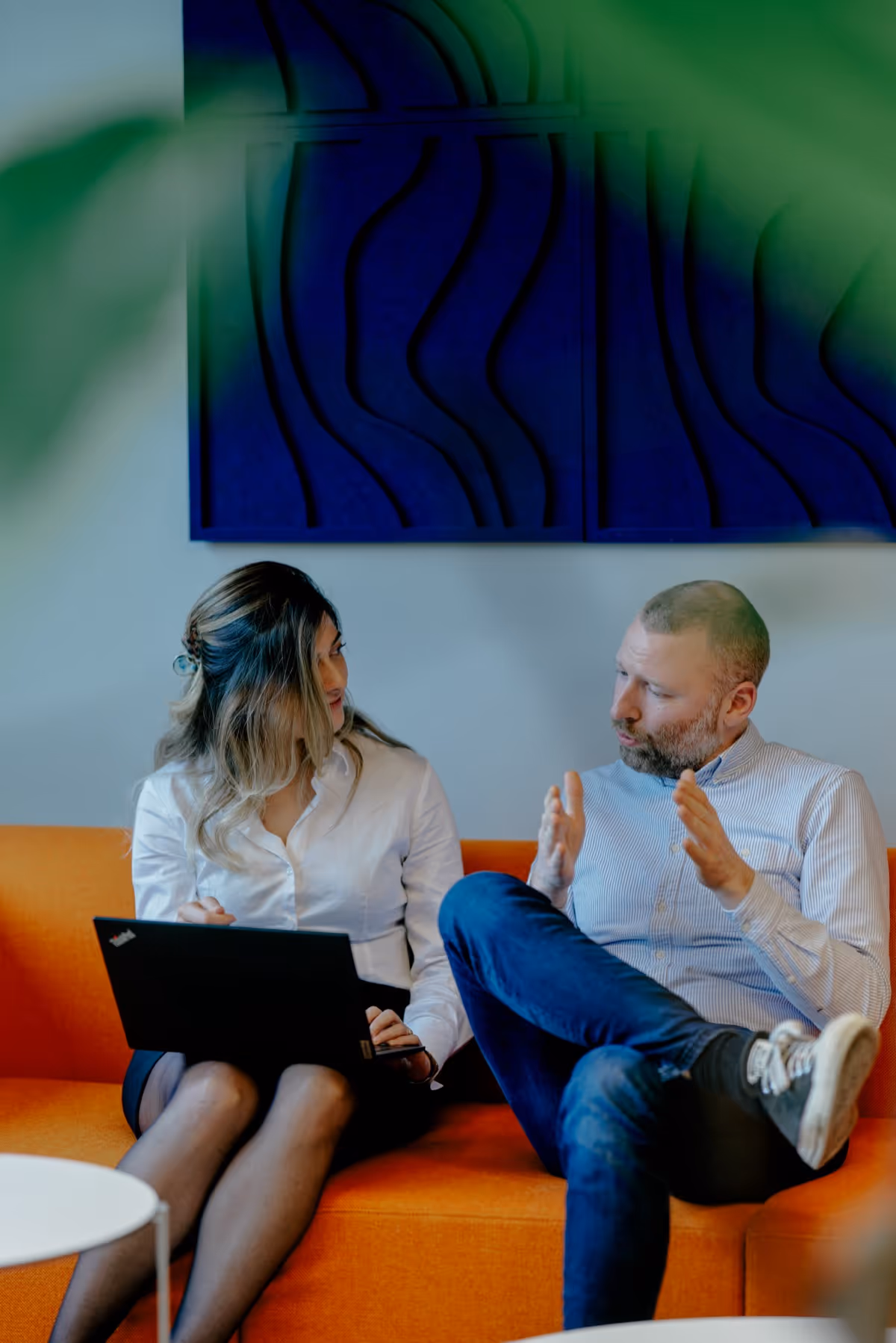 A man and a woman sitting on an orange couch, discussing with a laptop on the woman's lap.