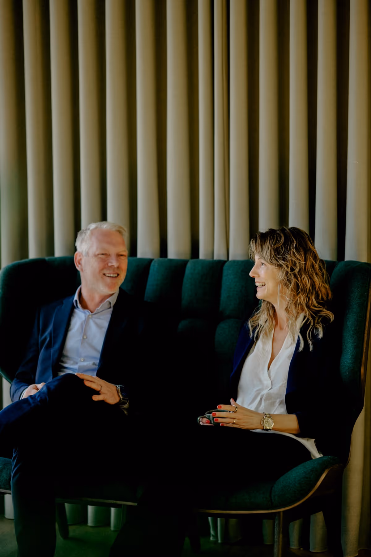 Two professionals sitting on a green couch, smiling and engaged in conversation against a backdrop of beige curtains.