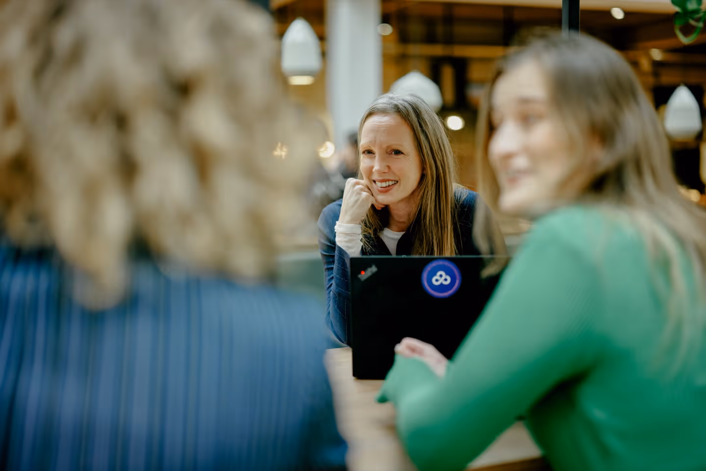 Three women seated at a wooden table, one woman facing the camera smiling with a laptop in front of her, the other two women blurred in the foreground.