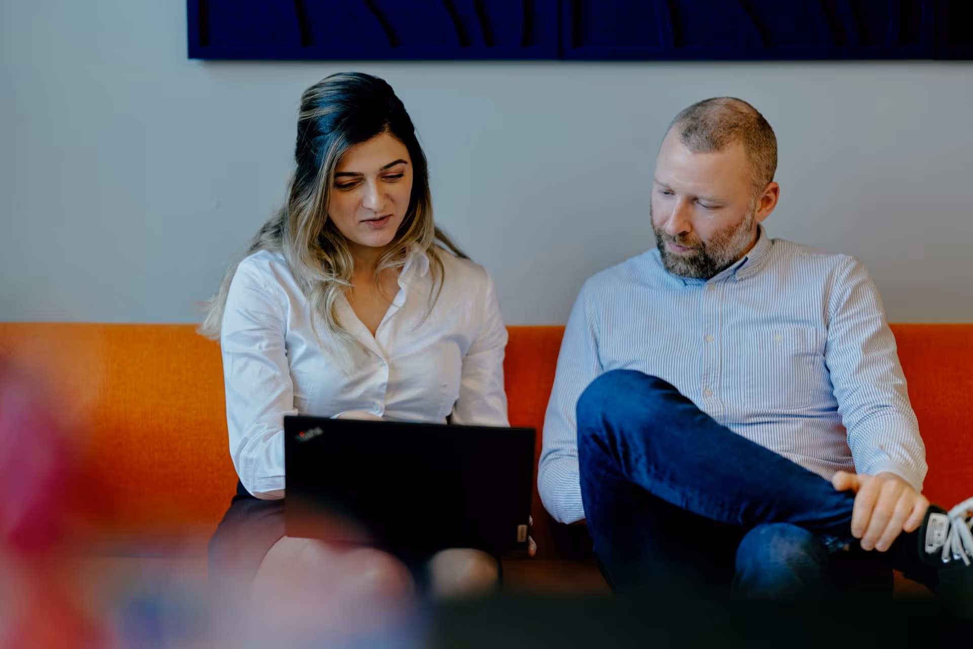 A woman and a man sitting on an orange couch looking at a laptop together.