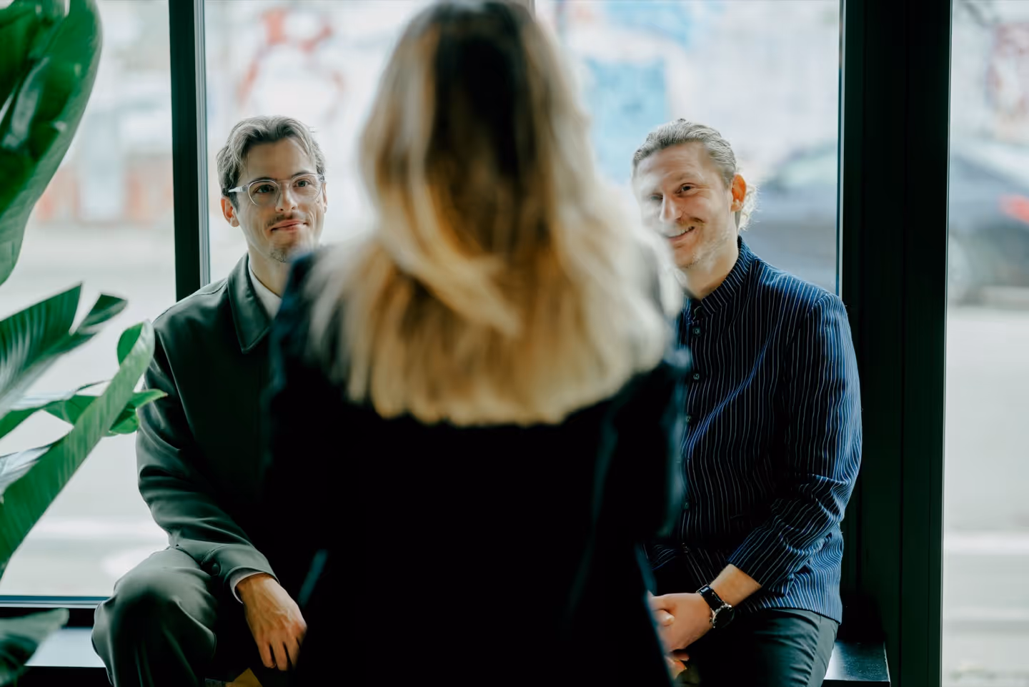 Two men sitting by a large window smiling and looking at a blonde woman standing with her back to the camera.