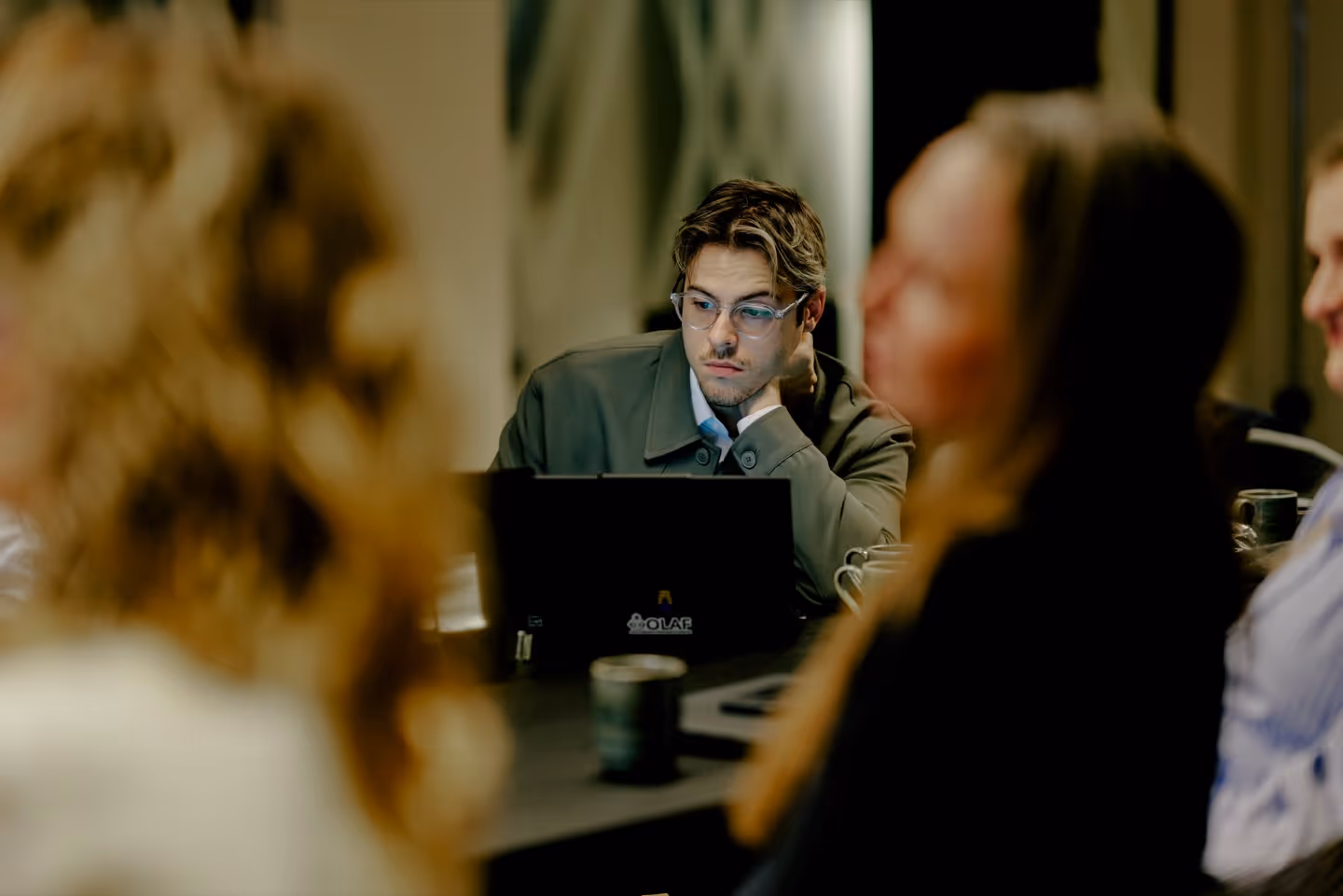 Young man wearing glasses and a green jacket looking thoughtfully at a laptop during a meeting with blurred colleagues in the foreground.
