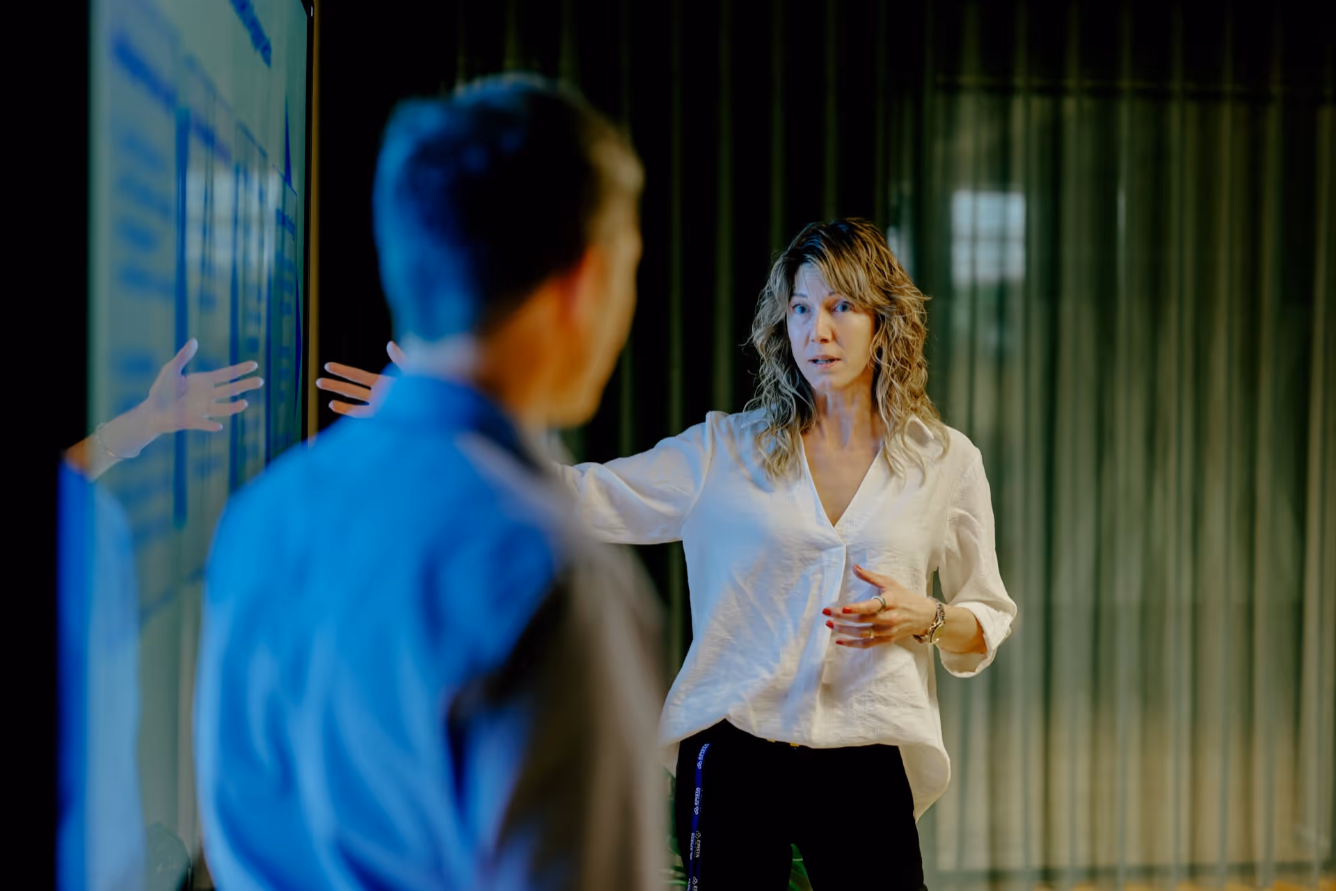 Woman in white shirt explaining information on a large screen to a man in a blue shirt in a dimly lit room.