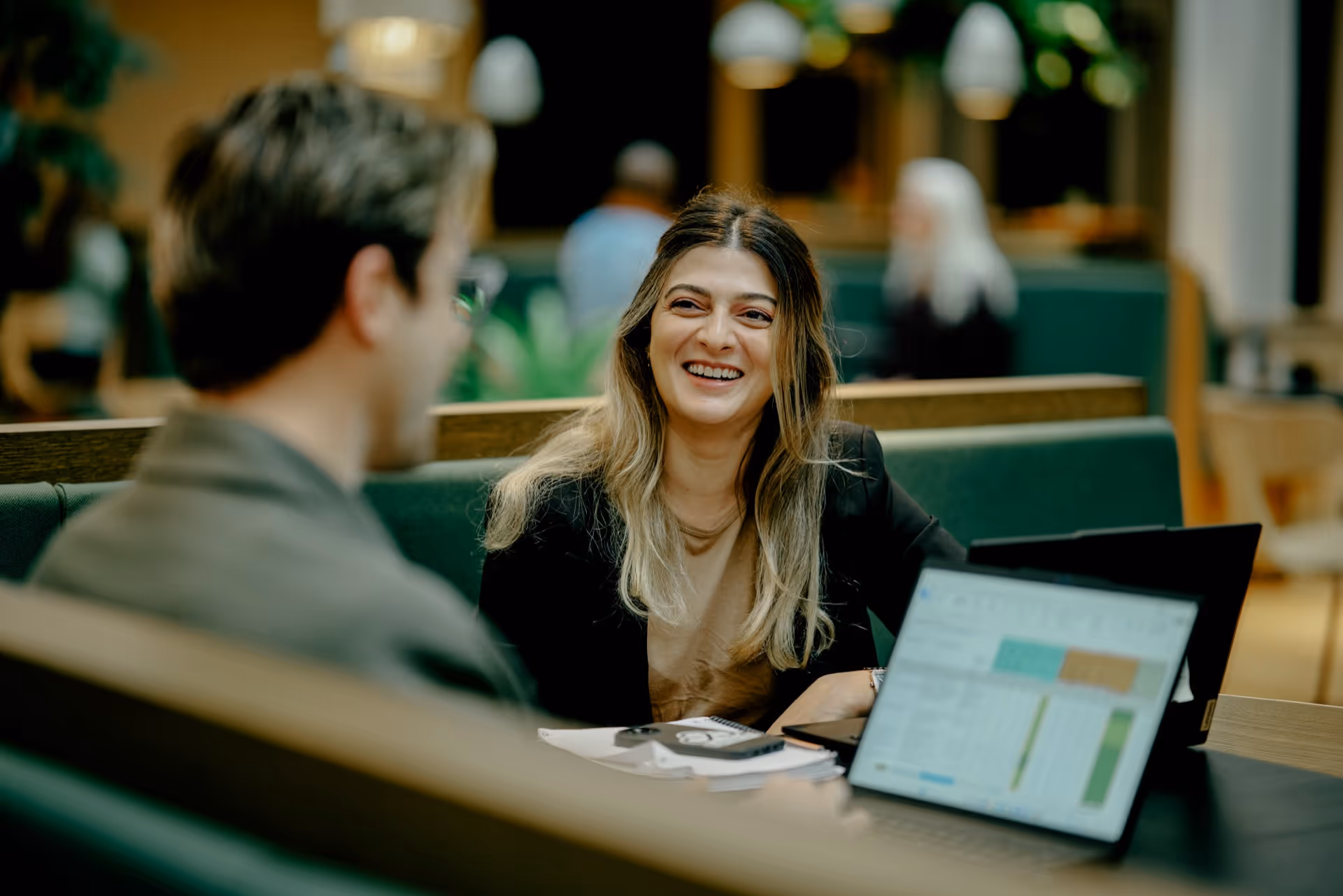 Smiling woman in a professional setting talking with a man, with a laptop and papers on the table.