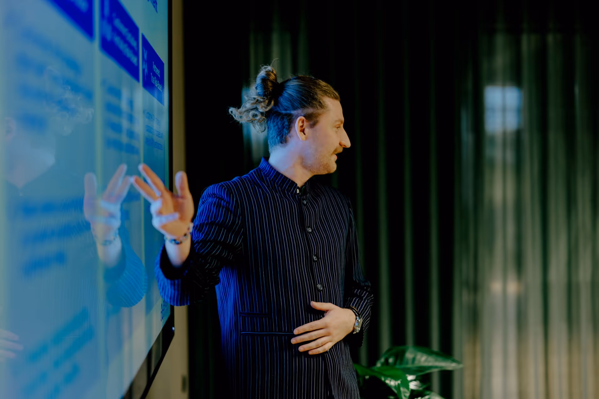 Man with a bun hairstyle giving a presentation and pointing at a large screen with charts.