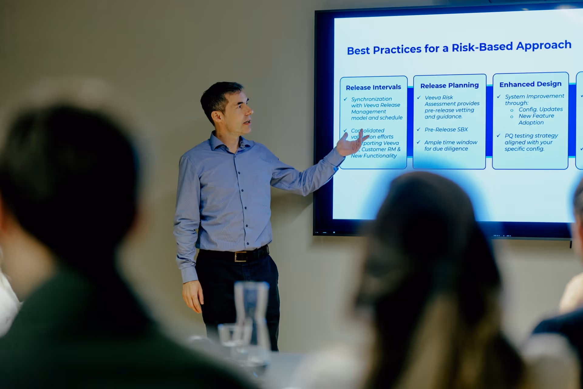 Man in a blue shirt giving a presentation pointing at a screen displaying 'Best Practices for a Risk-Based Approach' with release intervals, release planning, and enhanced design details.