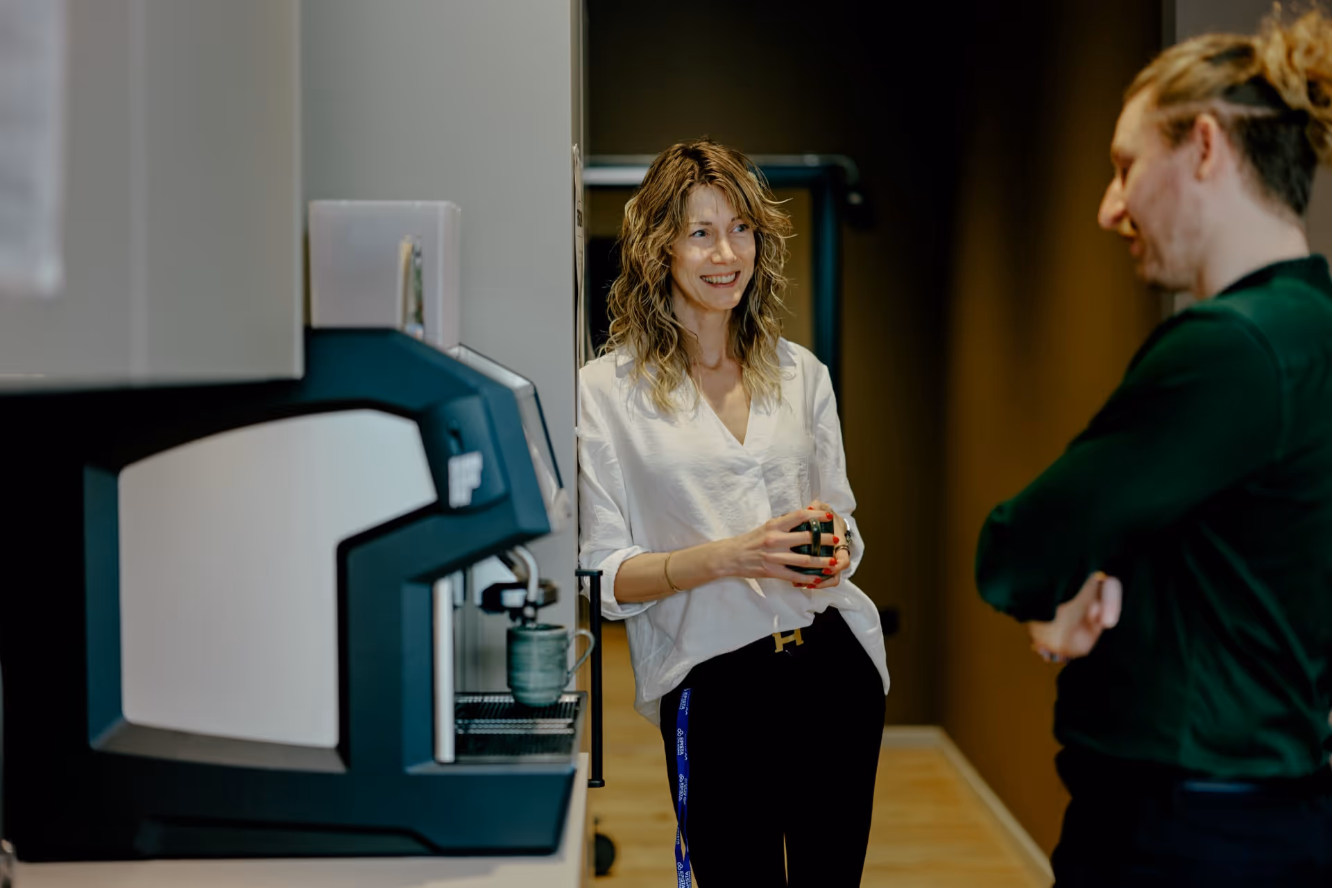 Two colleagues having a friendly conversation near a coffee machine in an office kitchen.